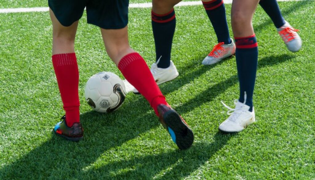 The legs of three people playing football. They have red and black football socks and white trainers