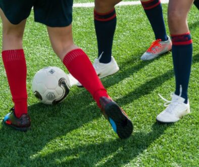 The legs of three people playing football. They have red and black football socks and white trainers
